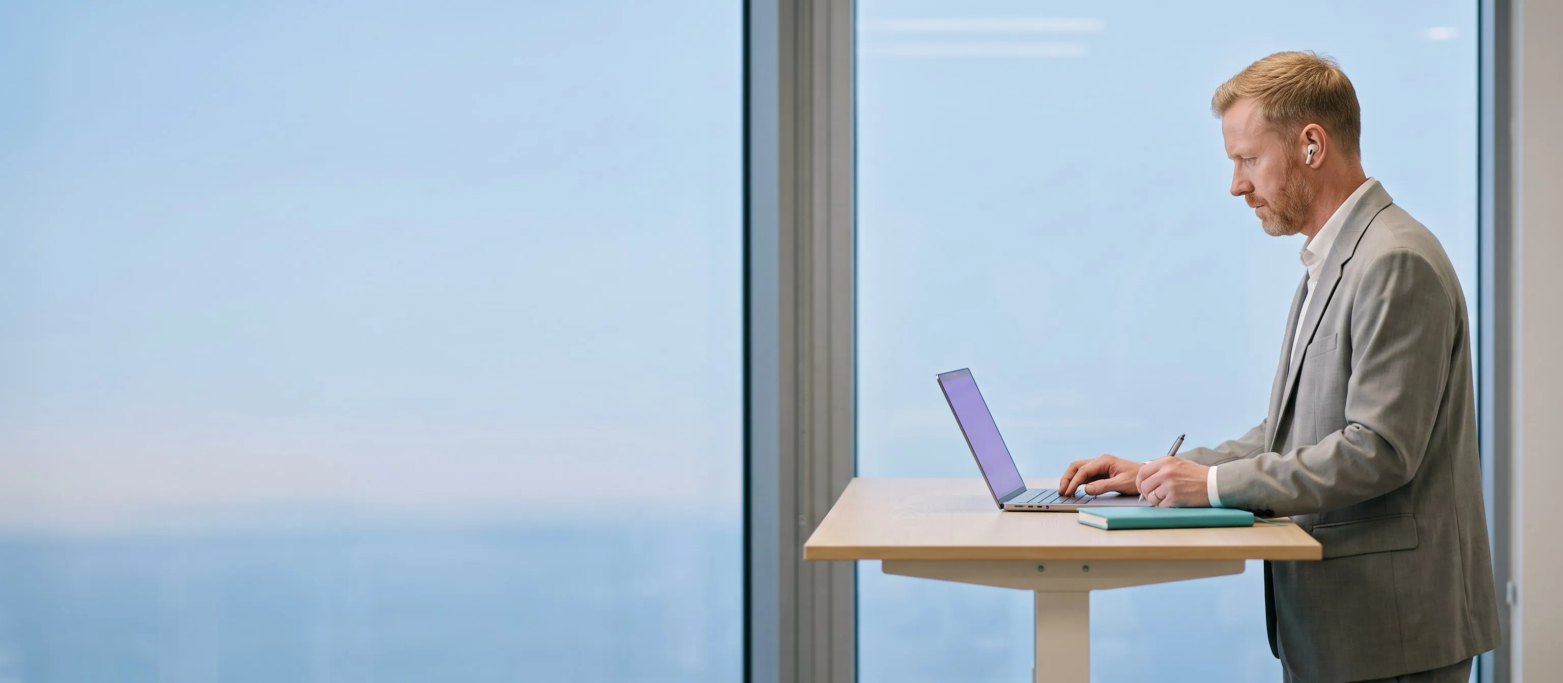 A businessman working on a laptop at a standing desk beside a tall office window